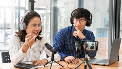 Asian woman radio hosts gesturing to microphone while interviewing a man guest in a studio while...