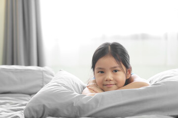 Asian child smile or kid girl happy lie down or sleep on soft pillow to comfortable on gray bed relax and white curtain with window light and stay home safe in morning to wake up or woke up at bedroom