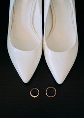 Fototapeta premium Gold wedding rings and white shoes of the bride on a dark background.