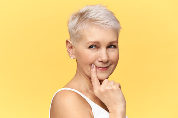 Hmm, let me think. Isolated image of attractive retired female with short haircut keeping fore finger on her lips, trying to guess something, having thoughtful suspicious facial expression