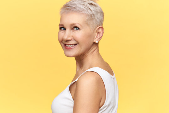 Isolated image of gorgeous charming middle aged woman with dyed short hair turning head, smiling happily, posing against blank yellow background with copy space for your advertising content