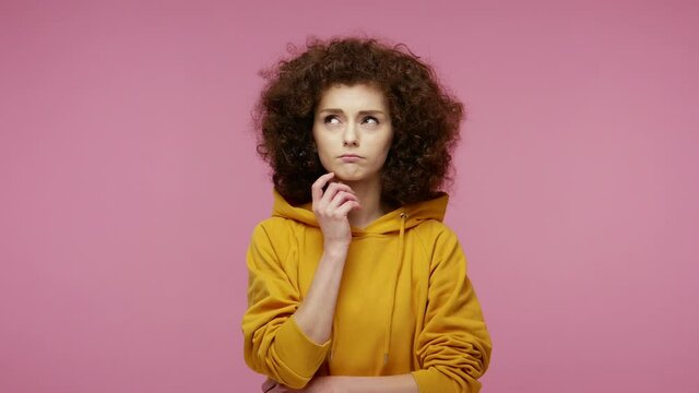 Thoughtful confused girl afro hairstyle in hoodie touching chin, scratching head while thinking over solution, having doubts about difficult choice. indoor studio shot isolated on pink background
