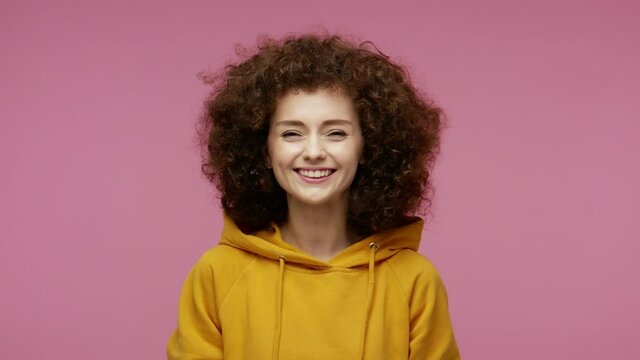 Carefree positive naughty girl afro hairstyle in hoodie sticking tongue out, making faces having fun in front of camera, grimacing expressing disobedience. studio shot isolated on pink background