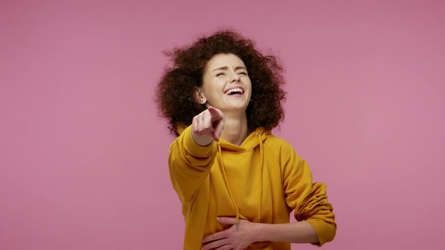 Hey you! Amused girl afro hairstyle in hoodie pointing finger to camera, laughing out loud, taunting making fun of ridiculous appearance, funny joke. indoor studio shot isolated on pink background