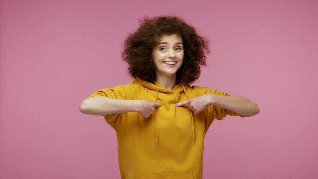 This is me! Happy excited girl afro hairstyle in hoodie pointing herself, looking around and bragging everyone about own success, proud of achievement. indoor studio shot isolated on pink background