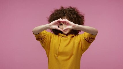 Be my Valentine! Cute happy girl afro hairstyle in hoodie making heart shape with hands and kissing air, demonstrating love affection, romantic feelings. indoor studio shot isolated on pink background