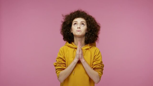 Please god, I'm begging! Upset girl afro hairstyle in hoodie appealing to heaven with pleading imploring eyes, keeping prayer gesture and asking help. indoor studio shot isolated on pink background