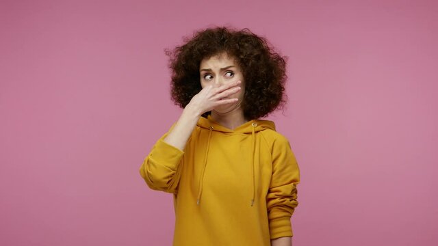 Confused displeased girl afro hairstyle in hoodie feeling disgust repulsion to bad smell, pinching her nose holding breath to avoid stink, fart gases. indoor studio shot isolated on pink background