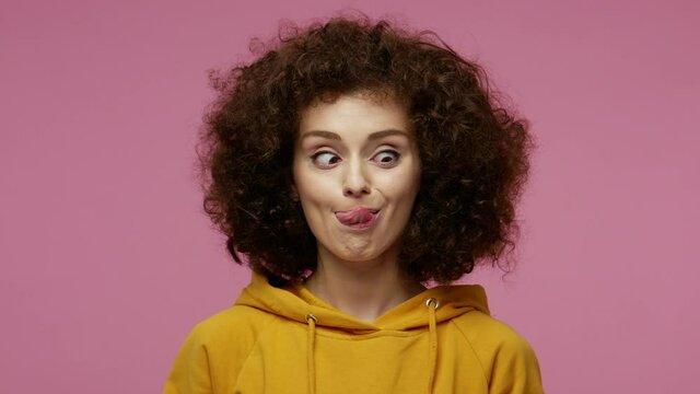 Funny amusing girl afro hairstyle in hoodie looking awkward with crossed eyes and puffed cheeks, fooling around making stupid brainless dumb expression. indoor studio shot isolated on pink background