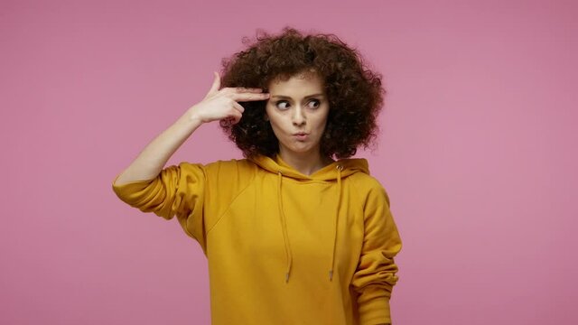 Suicide gesture. Depressed girl afro hairstyle in hoodie aiming finger gun to head, shooting killing herself, pretending to die with crossed eyes and tongue out. indoor studio shot isolated on pink