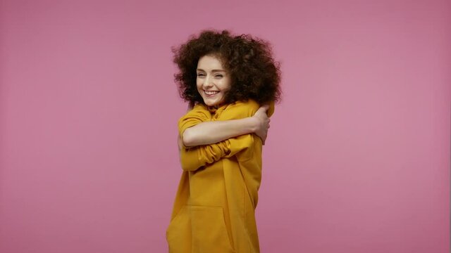 Adorable girl afro hairstyle in hoodie embracing herself, hugging with self-love and smiling from pleasure, narcissistic egoistic feelings, complacency concept. studio shot isolated on pink background