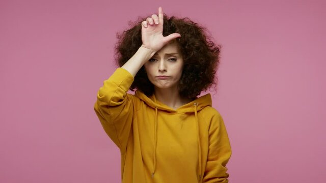 Unlucky sad girl afro hairstyle in hoodie showing loser or lame gesture, L finger sign on forehead, life troubles and unemployment concept, fired from job. studio shot isolated on pink background