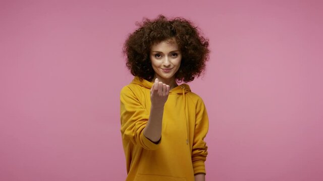 Come here, follow me! Beautiful girl afro hairstyle in hoodie making beckoning gesture with one finger, inviting to approach, looking playful flirting. indoor studio shot isolated on pink background
