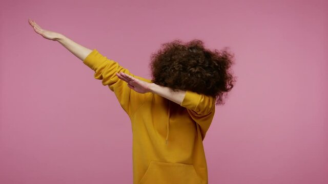 Trendy girl afro hairstyle in hoodie performing dab dance with serious calm face and making dabbing movement, with hands up, internet meme gesture. indoor studio shot isolated on pink background