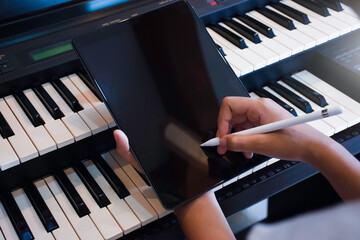 Woman hand holding stylus pencil and writing on black blank screen tablet computer pc with piano background in music room.