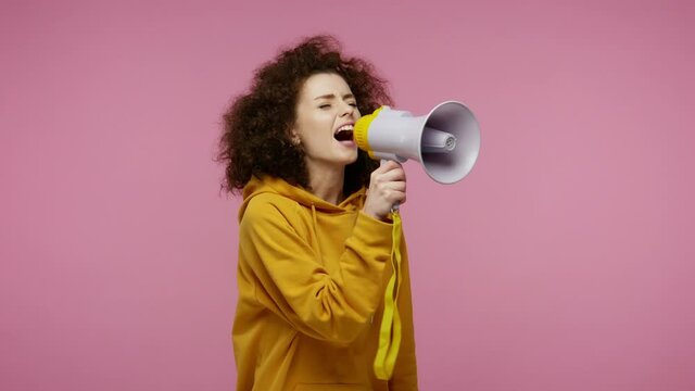 Funny girl afro hairstyle in hoodie talking with megaphone, proclaiming news, loudly announcing advertisement, warning using loudspeaker to shout speech. studio shot isolated on pink background