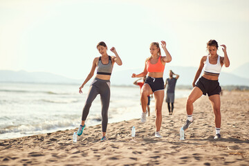group of three women exercise on a beach with two men walking in the background