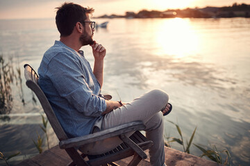 beardy guy sitting alone on a river coast, enjoying the sunset, thinking