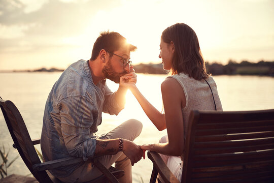 beardy man holding and kissing hand of a woman beside river at sunset