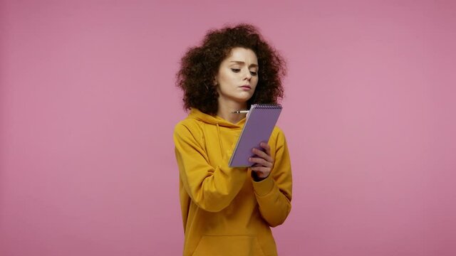 Thoughtful creative girl afro hairstyle in hoodie thinking over future business plan, taking notes in paper notebook, writing creative idea and smiling inspired happy. indoor studio shot isolated