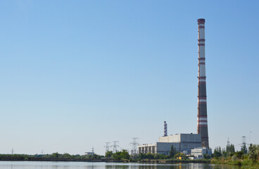 view from afar of the buildings and the pipe of the power plant. lake water and blue sky are also visible