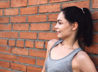 Obraz na płótnie Canvas Close-up portrait of a smiling woman. Young woman rests after jogging against a red brick wall. Caucasian female fitness model working out in the morning. Female athlete resting after training session