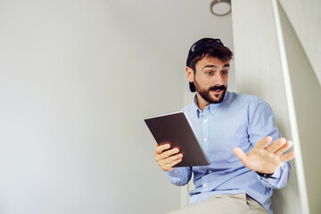 Young handsome businessman dressed business casual sitting in office and using tablet while talking to colleague.