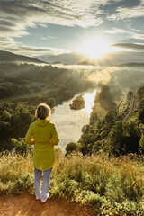Naklejka premium Happy girl in a yellow jacket enjoying sunrise on cliff.Beautiful view of morning landscape with fog and sun rays.Hiking in the countryside.Wanderlust and travel concept.Atmospheric moment copy space