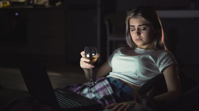 Upset woman drinking alcohol, watching a sad movie on TV at home, sitting on a sofa alone. Unhappy young people, negative emotions and television concept.