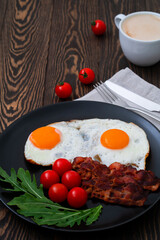 Tasty breakfast with fried eggs, crispy bacon, cherry tomatoes and arugula served on the black plate on the wooden table with cup of coffee and knife and fork on the napkin. 