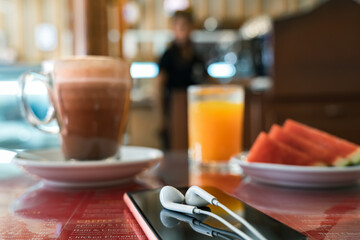Cup of coffee, smartphone with headphones, freshly squeezed cold orange juice and plate with slices of watermelon on a cafe table