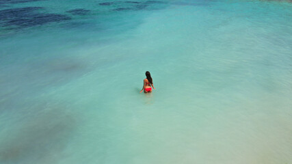 Aerial view of young beautiful woman in red bikini swimming in light blue azure turquoise water of sea, relaxing in ocean. Summer beach vacation. Malibu lifeguard Maldives Melasti beach Bali Indonesia