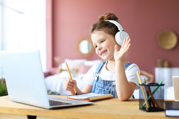 Distance learning. Cheerful little girl girl in headphones using laptop studying through online...