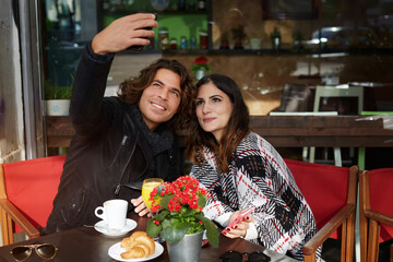 Couple in love breakfast with warm coffee and delicious dessert in sidewalk cafe, attractive man and woman taking picture of themselves with mobile phone camera during vacation holidays in autumn