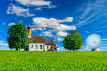 Kleine Kirche und die Parabolantenne einer Erdfunkstation zwischen grünen Feldern und Bäumen.