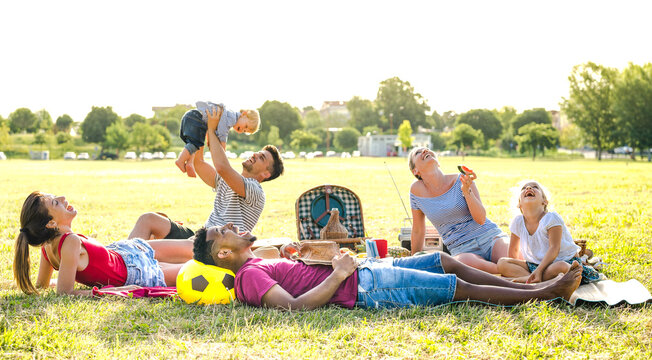 Young multiracial families having fun playing with kids at pic nic garden party - Multiethnic joy and love concept with mixed race people together with children at park barbecue - Warm bright filter