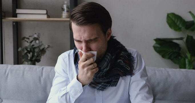 Young man sitting on couch in living room cover mouth with paper tissue coughs holds his hand in lung area feels unhealthy, male got sick, seasonal flu. Symptom, sign of corona virus covid19 concept