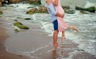 The guy holds the girl in his arms on the beach in the water barefoot in the wet sand. Man and woman on the seashore in waves with foam and stones in a water, tender embrace, love concept