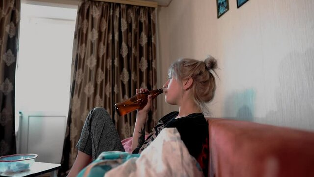 The girl drinks beer and watches football. Her boyfriend cleans the room. Henpecked in the house.