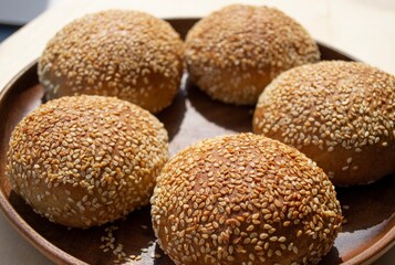 Art composition in the rays of the bright daylight. Homemade round rye buns with sesame seeds on a wooden plate (focus on the first two, on the distant buns, the focus is specially blurred)