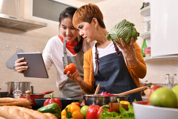Happy family of mother and daughter using digital tablet and cooking in kitchen making healthy food together feeling fun