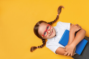 Cute little girl schoolgirl in glasses with a book on a yellow background. Top view