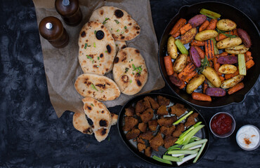 Roasted potatoes and carrots, celery and herbs in cast iron pan on dark table, copy space
