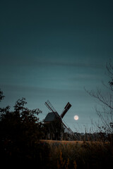 Landscape countryside view of a wodden windmill with full moon rise and blue night sky with fields....