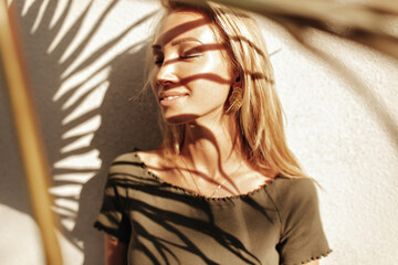 Pretty blonde long-haired girl in dark stylish top and with gold earrings smiles with closed eyes and poses near white wall with palm leaf shadow.