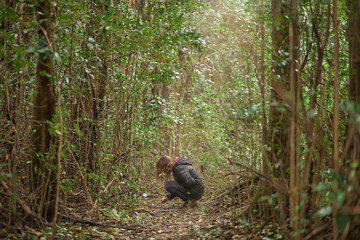 Blond woman crouched in the forest observing tree leaves on the ground illuminated by a light beam