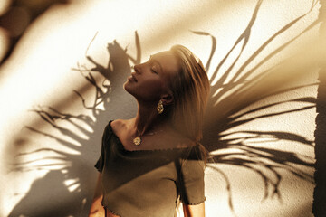 Attractive blonde woman in trendy dark green t-shirt poses with closed eyes near wall with palm...