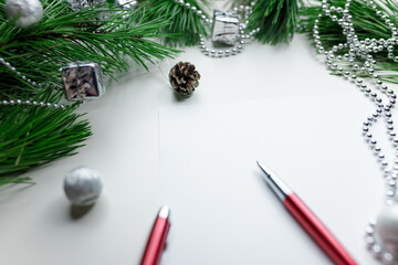 Christmas holiday background - decoration on a white table background with blank white sheet and red pen. New year pine and silver Christmas balls. Top view. Place for text.