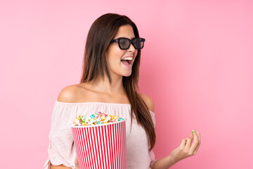 Teenager girl over isolated pink background with 3d glasses and holding a big bucket of popcorns while looking side