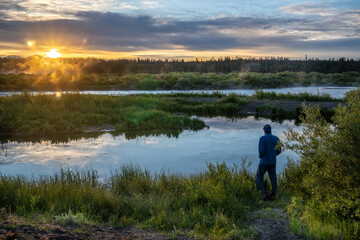 Man standing medium watching the sunrise over the mountains and under clouds with reflection in the nearby river, Madison River, Montana